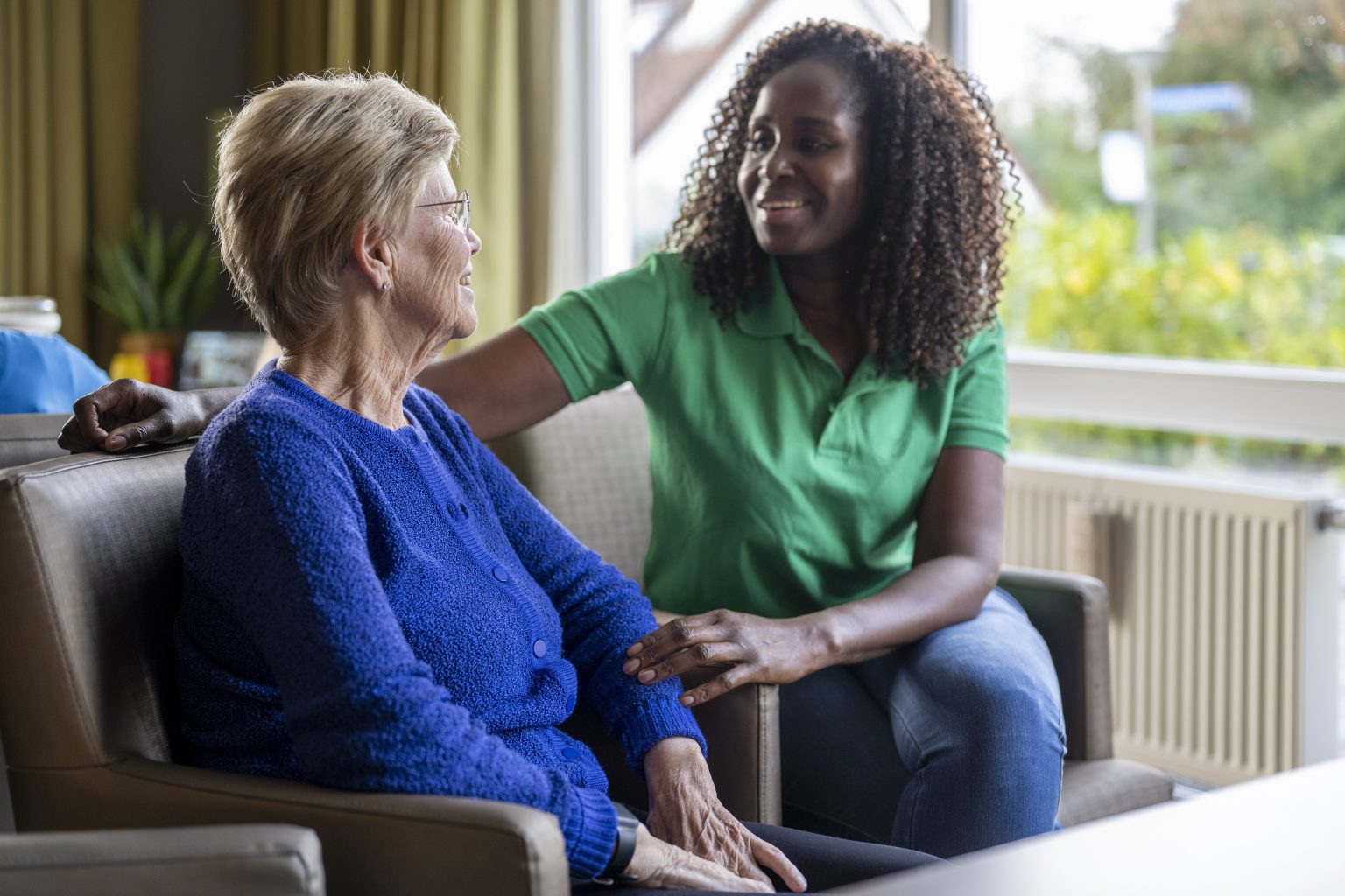 Alltagsbegleiterin sitzt lächelnd neben einer Seniorin im Wohnzimmer