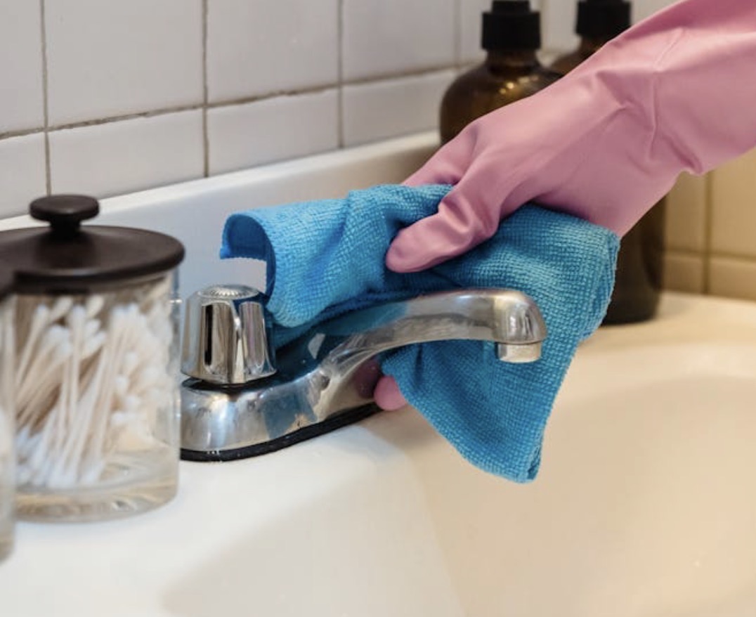 Behandschuhte Hand reinigt einen Badezimmer-Wasserhahn mit einem Tuch